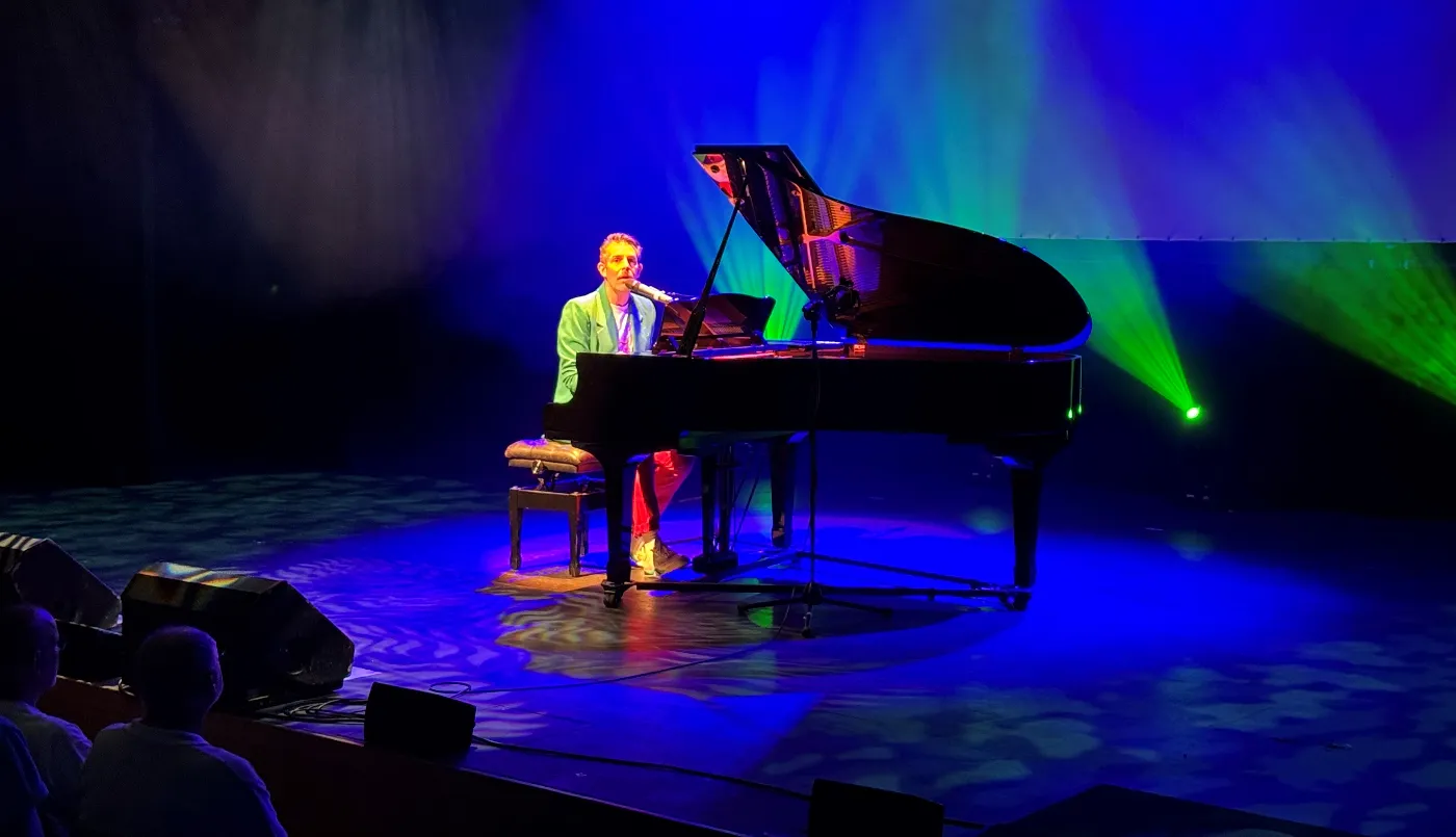 A man on stage plays a grand piano and sings. The room is dark and the stage is illuminated with blue and green stage lights