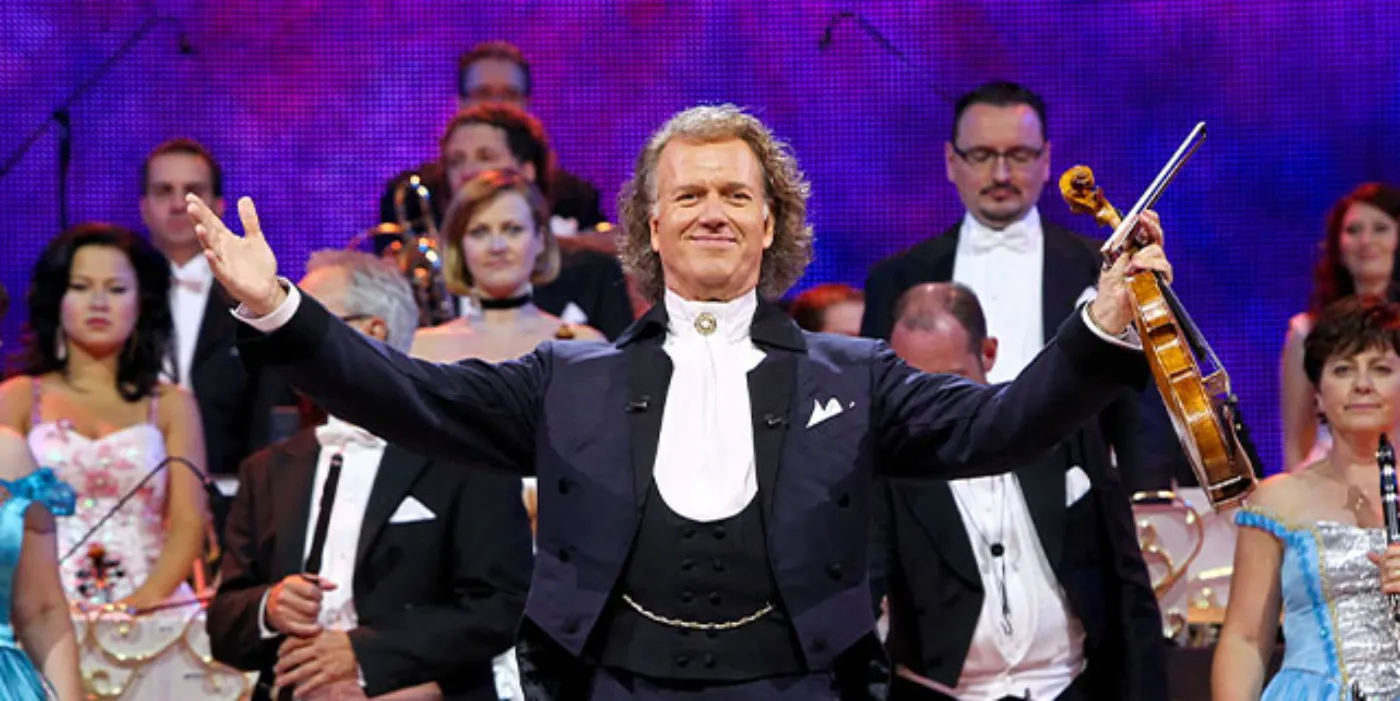 A violinist in a tuxedo faces the crowd looking happy with his arms out-stretched. The orchestra can be seen standing behind him in the background